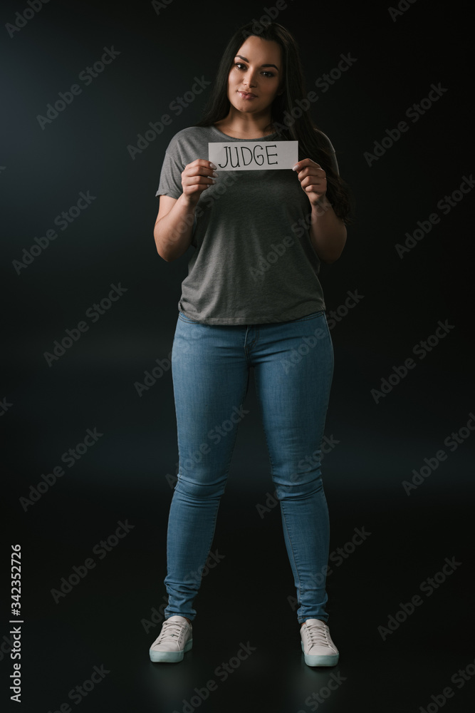 Full length view of smiling plus size girl holding card with word judge on black background Full length view of smiling plus size girl holding card with word judge on black background