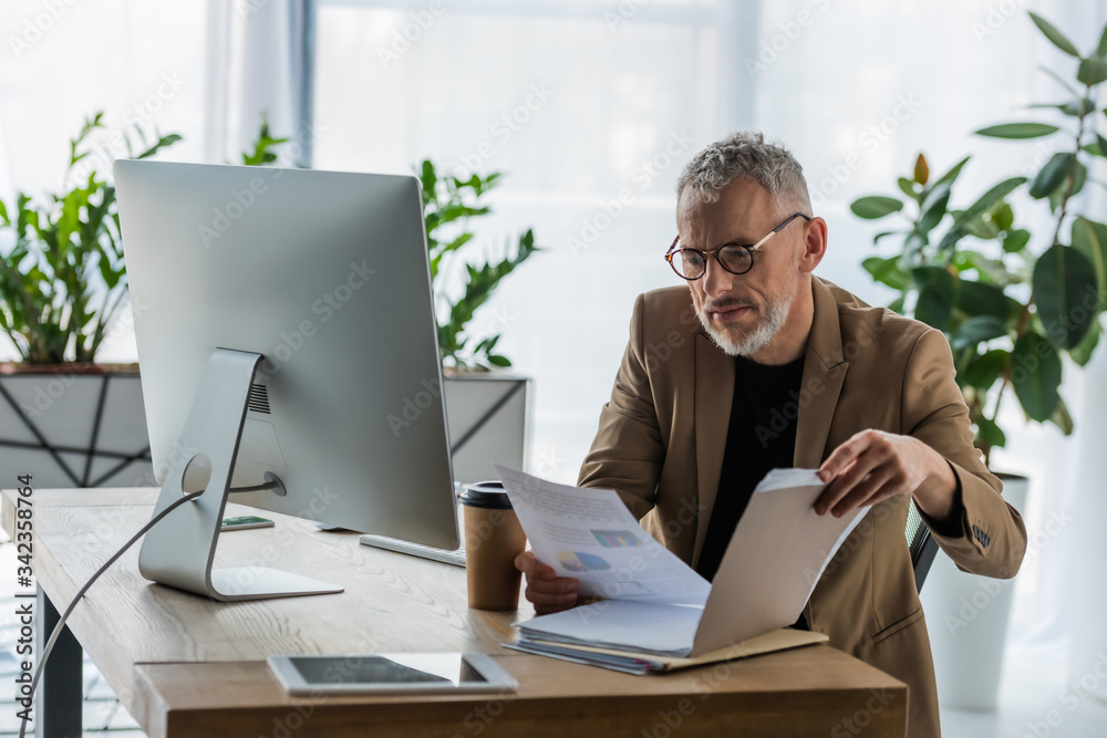 selective focus of bearded businessman in glasses looking at document near coffee to go and computer monitor in office selective focus of bearded businessman in glasses looking at document near coffee to go and computer monitor in office