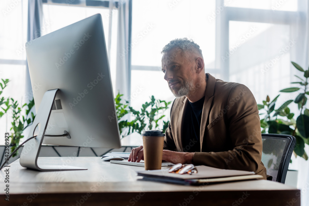 selective focus of bearded businessman working near coffee to go and computer monitor in office selective focus of bearded businessman working near coffee to go and computer monitor in office