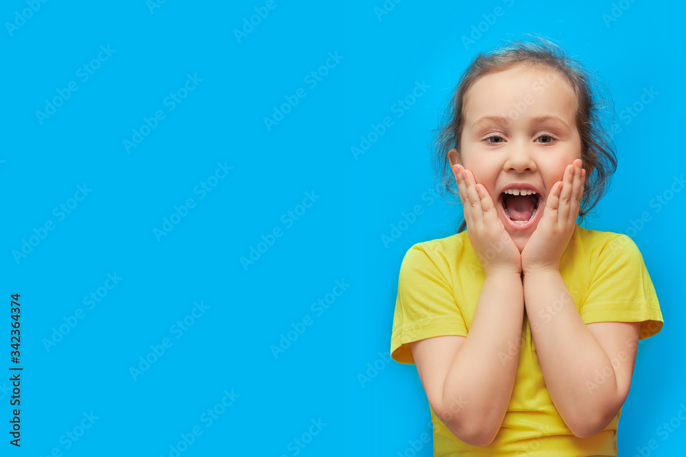 Emotional portrait of a preschool girl on a blue background. A little girl with curly hair opens her mouth wide and screams with delight. He holds his hands to his face. Layout. copy space Emotional portrait of a preschool girl on a blue background. A little girl with curly hair opens her mouth wide and screams with delight. He holds his hands to his face. Layout. copy space