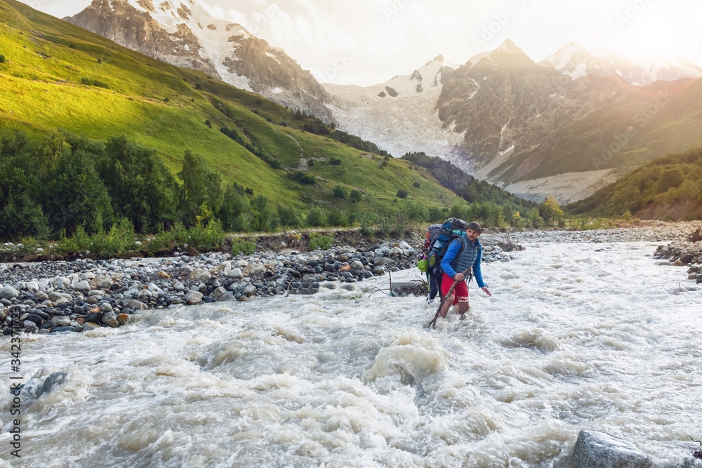 A backpacker with a backpacker goes across a swift mountain river. Crossing the mountain river wading. The mountain river flows from the Adisha glacier in the Caucasus. The tourist crosses the river. A backpacker with a backpacker goes across a swift mountain river. Crossing the mountain river wading. The mountain river flows from the Adisha glacier in the Caucasus. The tourist crosses the river.
