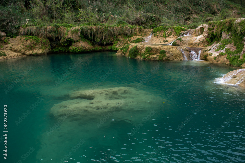 Poza de agua color turquesa con formaciones de piedra caliza debajo del agua rodeado de caídas de agua en la roca cubierta de helechos 