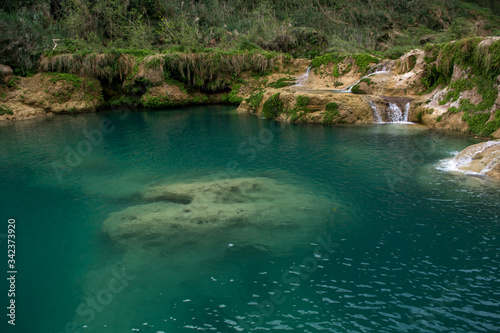 Poza de agua color turquesa con formaciones de piedra caliza debajo del agua rodeado de caídas de agua en la roca cubierta de helechos 
