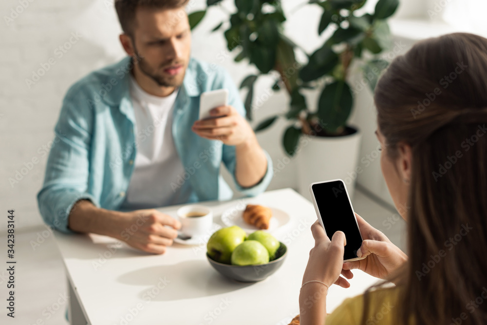 Selective focus of woman using smartphone near boyfriend chatting during breakfast Selective focus of woman using smartphone near boyfriend chatting during breakfast