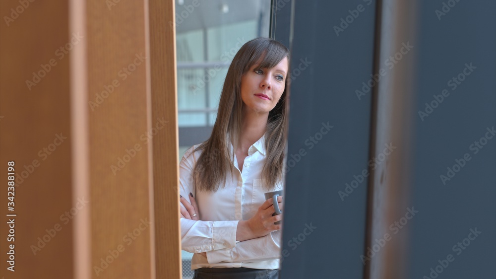 Businesswoman taking break in office drinking coffee standing in window and daydreaming, thinking.