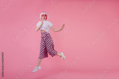 Full-length portrait of white adorable girl in peruke jumping with smile. Indoor photo of spectacular female model in stylish striped pants dancing in studio.