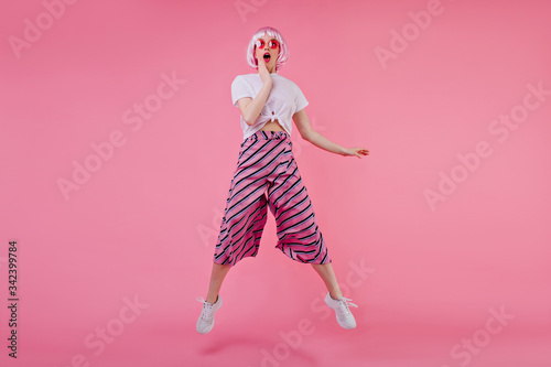 Full-length portrait of surprised happy girl in peruke jumping on bright background. Beautiful european female model having fun during photoshoot in periwig.