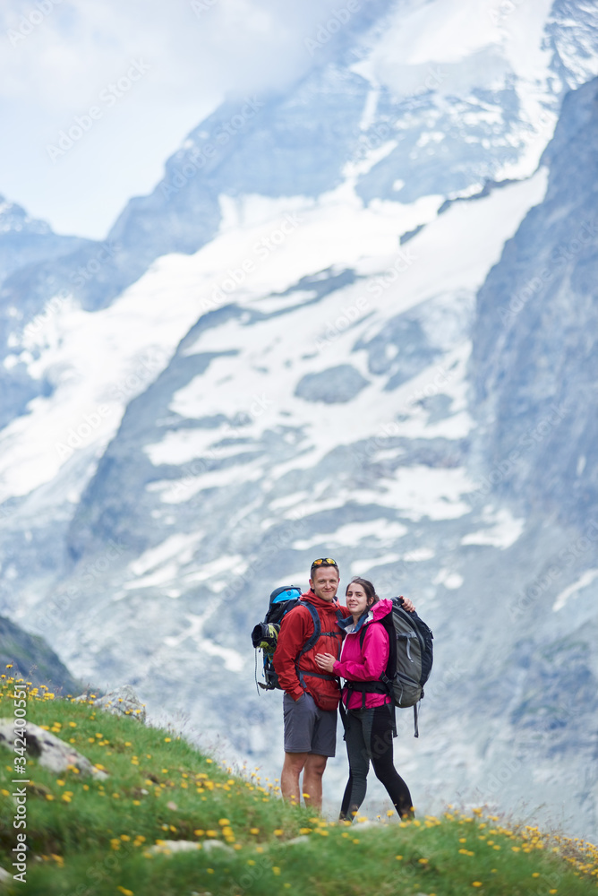 Portrait of couple of tourists travelling in Swiss Alps, man and woman standing on green meadow full of flowers on a breathtaking background with amazingly beautiful rocky mountains
