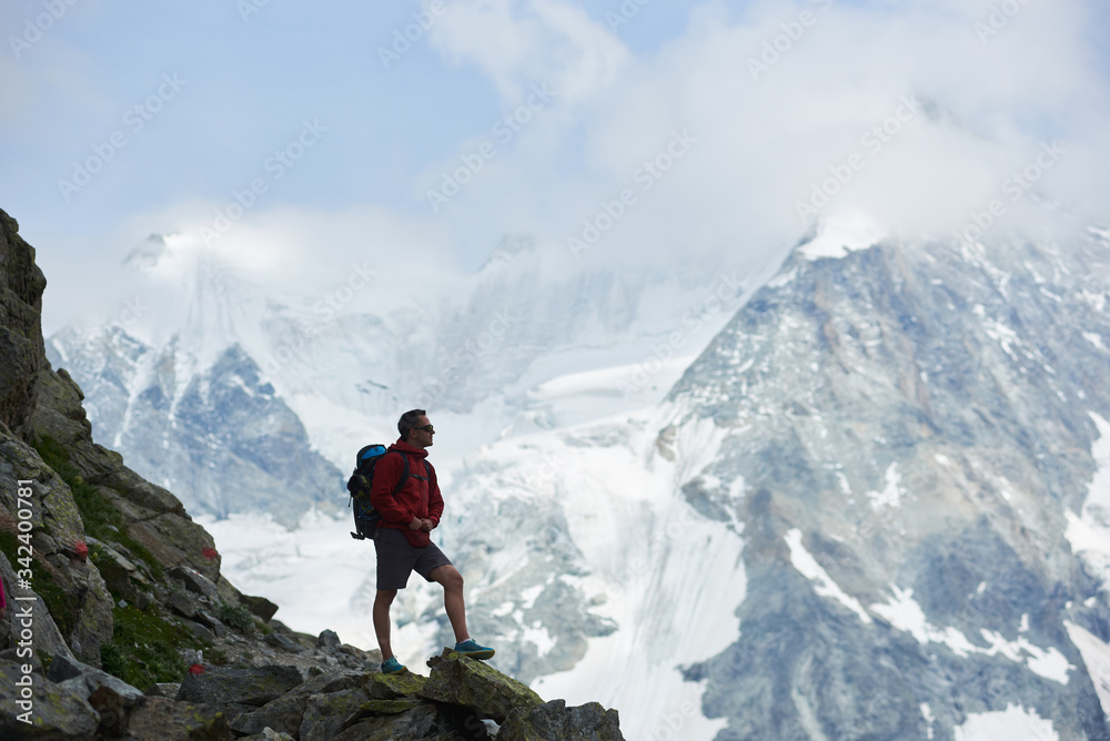 Side view of tourist with backpack standing on big stone, looking at beautiful mountains with snow. Trekking, mountain hiking, man reaching peak. Wild nature with amazing views. Sport tourism in Alps.