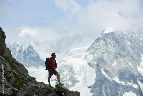Side view of tourist with backpack standing on big stone, looking at beautiful mountains with snow. Trekking, mountain hiking, man reaching peak. Wild nature with amazing views. Sport tourism in Alps. © anatoliy_gleb