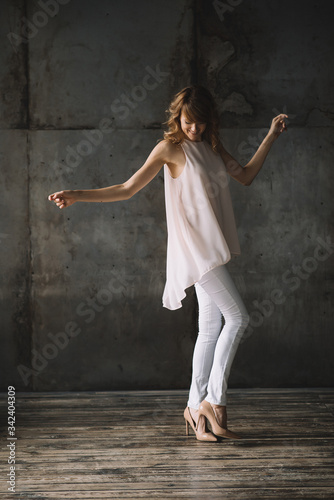 Beautiful young slender blond woman with curly hair in white trousers, a blouse and shoes in a loft studio is dancing. Soft selective focus.