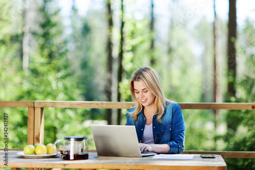 Blonde young woman working on laptop computer sitting at table on wooden balcony of country house