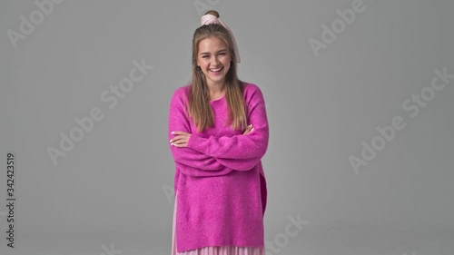 A beautiful young woman is laughing while standing with crossed hands isolated over grey background in studio photo