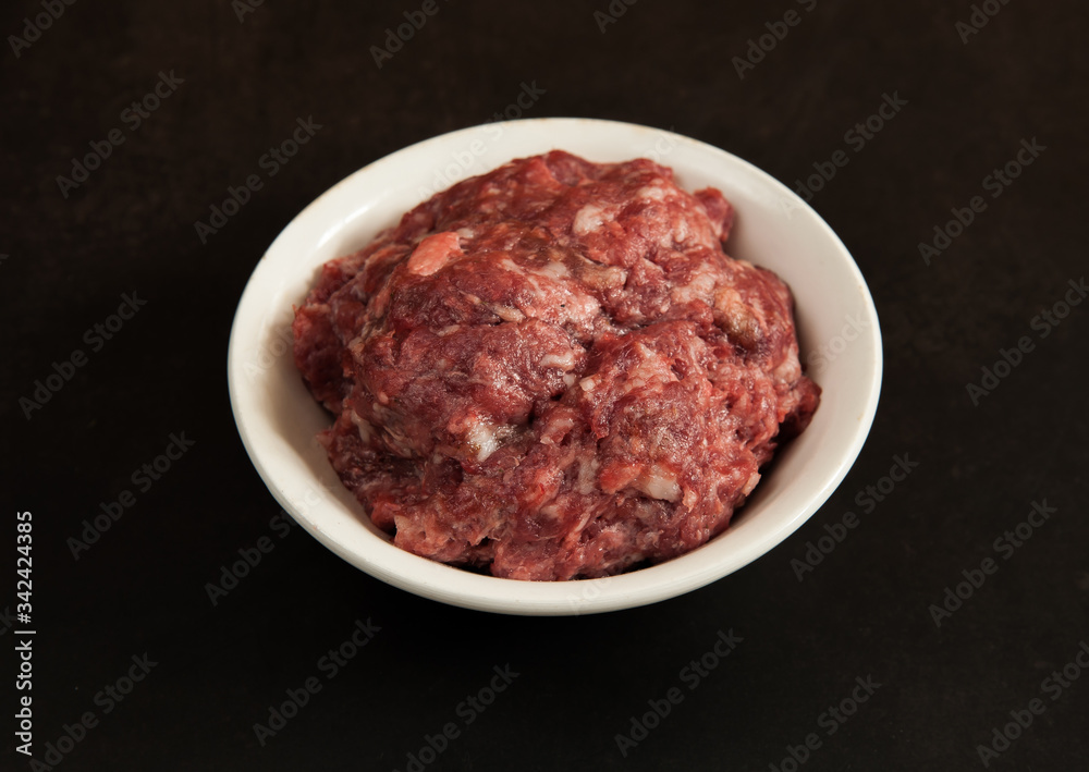 Raw fresh home-made ground beef in a white bowl close-up on a dark concrete background. Selective focus.