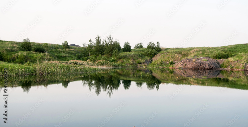 Lake in a coal pit with clear water at sunset. Reflection in the water of a coal mountain, hill, trees, reeds.The fuel industry produces coal in an open way.