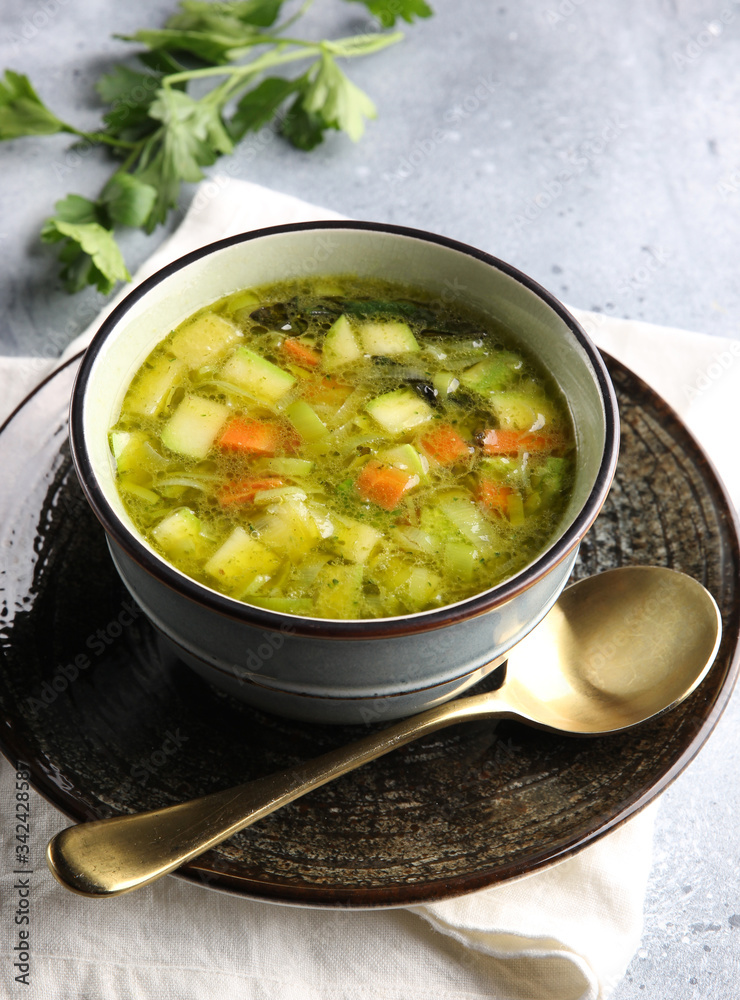 Lean vegetable soup with potatoes, zucchini, cabbage, carrots and herbs in a deep bowl with a spoon on a light grey background. Fresh parsley. Background image, copy space Lean vegetable soup with potatoes, zucchini, cabbage, carrots and herbs in a deep bowl with a spoon on a light grey background. Fresh parsley. Background image, copy space