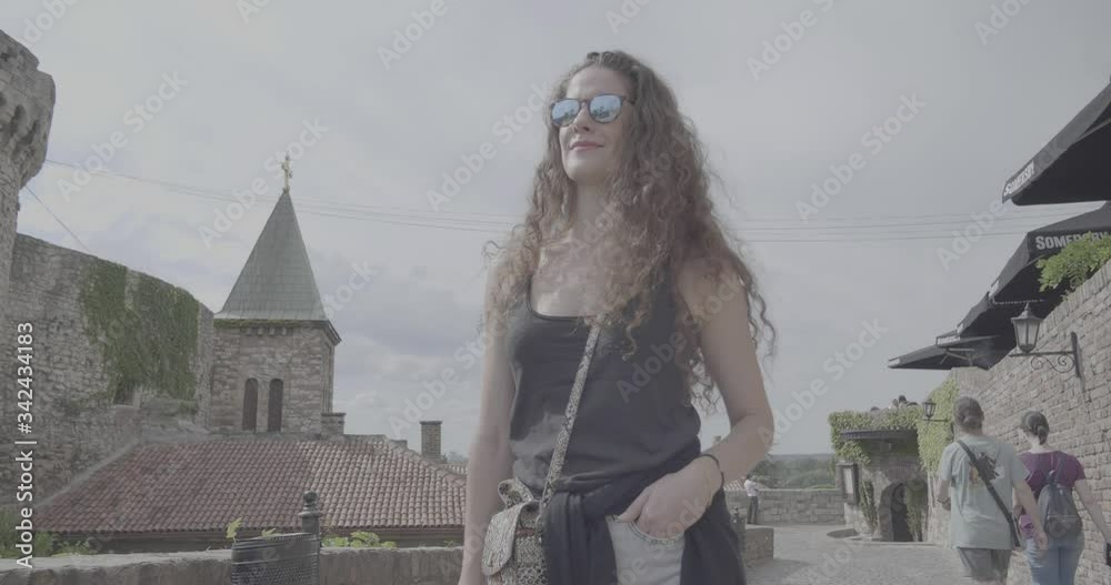 Young woman walking in the city, ancient wall background. Beautiful fashion model face with sunglasses, travel and explore the old town fortress in Belgrade Serbia, s-log ungraded