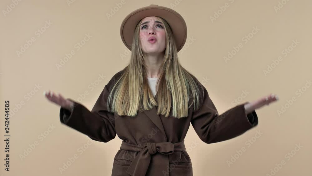 A displeased woman wearing hat and coat is raising her hands and screaming out isolated over the beige background in studio