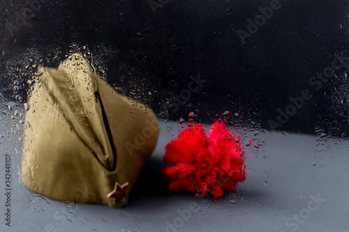 Window with rain drops. Forage,garisson cap of World War II soldier with red star,carnation flower.Memory of 75 years of victory day on May 9 of Soviet Union in Great Patriotic War.Dark background photo