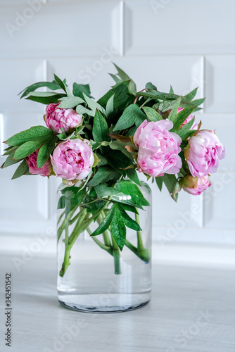 Bouquet of pink peonies in glass vase against white background and wooden table. Mothers day card. Beautiful peony flower for catalog or online store. Floral shop and delivery concept. Copy space.