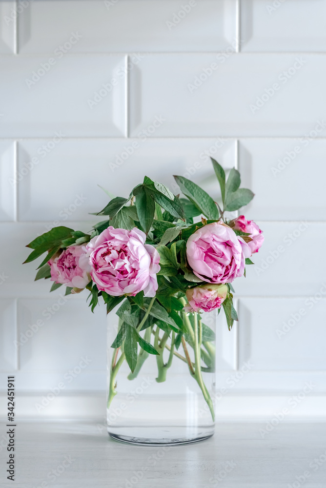 Bouquet of pink peonies in glass vase against white background and wooden table. Mothers day card. Beautiful peony flower for catalog or online store. Floral shop and delivery concept. Copy space.