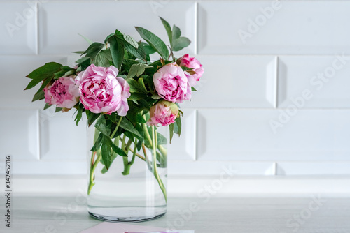 Bouquet of pink peonies in glass vase against white background and wooden table. Mothers day card. Beautiful peony flower for catalog or online store. Floral shop and delivery concept. Copy space.