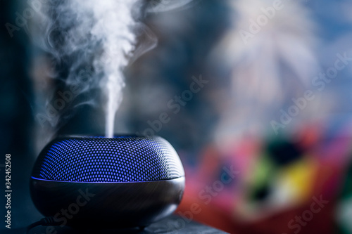 Aromatherapy oil diffuser electronic device with blue LED light from which steam comes out against a colored blurry background. Aromatherapy humidifier based on water and herbal essential oils.