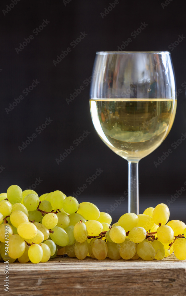 White wine in wineglass next to a bunch of grapes on a wooden table. Vertical portrait of alcohol drink and fruit on black background.