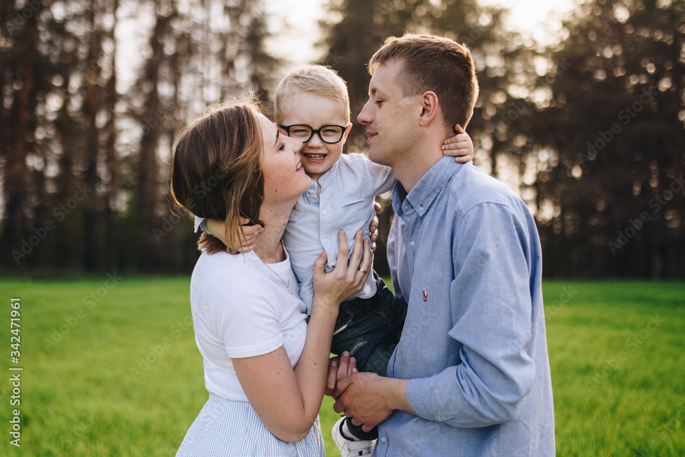 Fototapeta premium Family in nature. Picnic in the forest, in the meadow. Green grass. Blue clothes. Mom, dad, son with glasses. Boy with blond hair. Joy. Parents play with child. Together. Picnic basket. Food, blanket