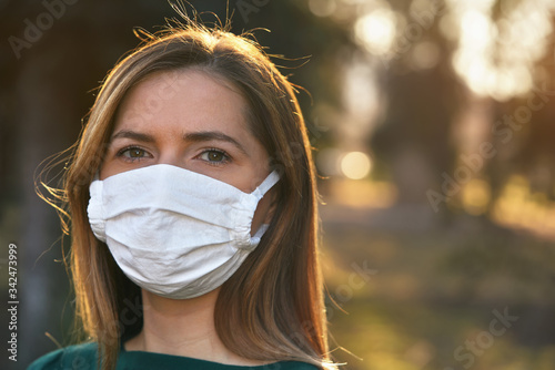Young woman wearing white cotton virus mouth nose mask, blurred park and trees in background, closeup face portrait