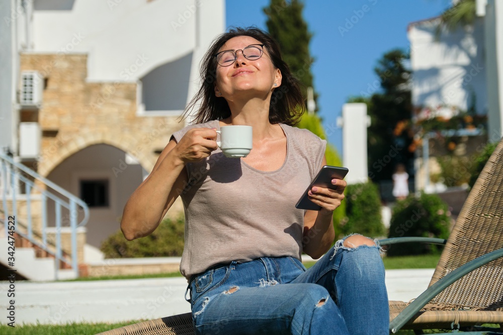 Mature woman relaxing in resort, sitting in chair with cup of coffee and smartphone