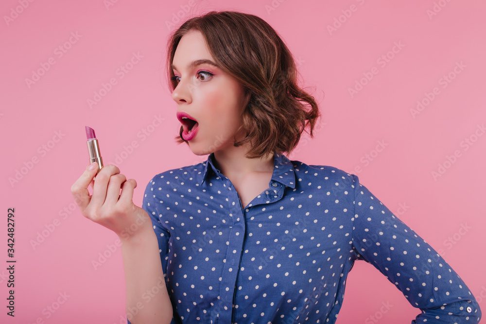 Funny lovely girl with wavy hairstyle looking at lipstick. Indoor photo of elegant female model in blue blouse doing makeup on pink background. Funny lovely girl with wavy hairstyle looking at lipstick. Indoor photo of elegant female model in blue blouse doing makeup on pink background.