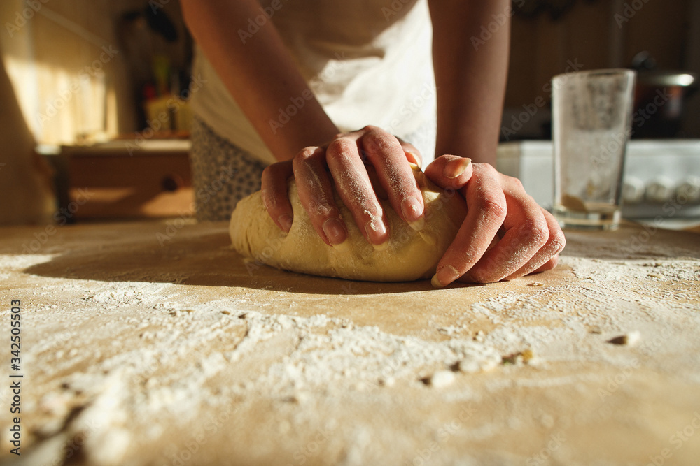 Woman's hands knead dough on a table in warm sunlight.