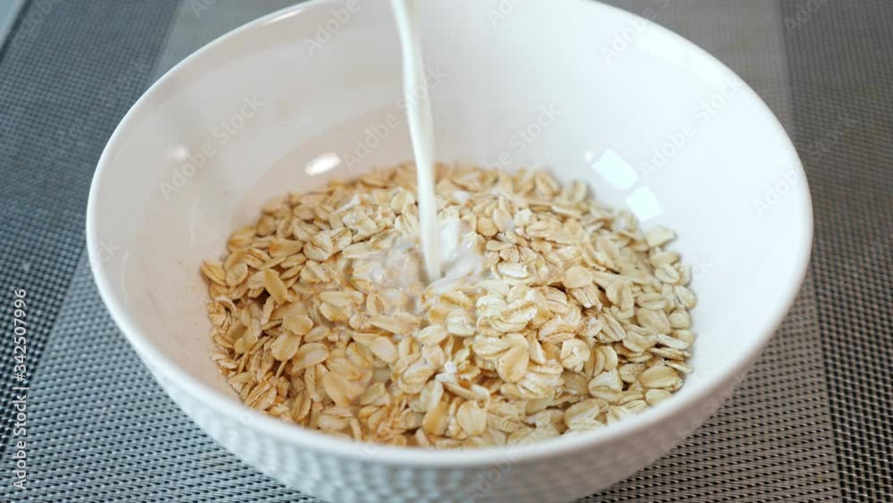 person pours fresh milk into ceramic bowl with tasty porridge cereals on grey table in hotel restaurant extreme close view person pours fresh milk into ceramic bowl with tasty porridge cereals on grey table in hotel restaurant extreme close view
