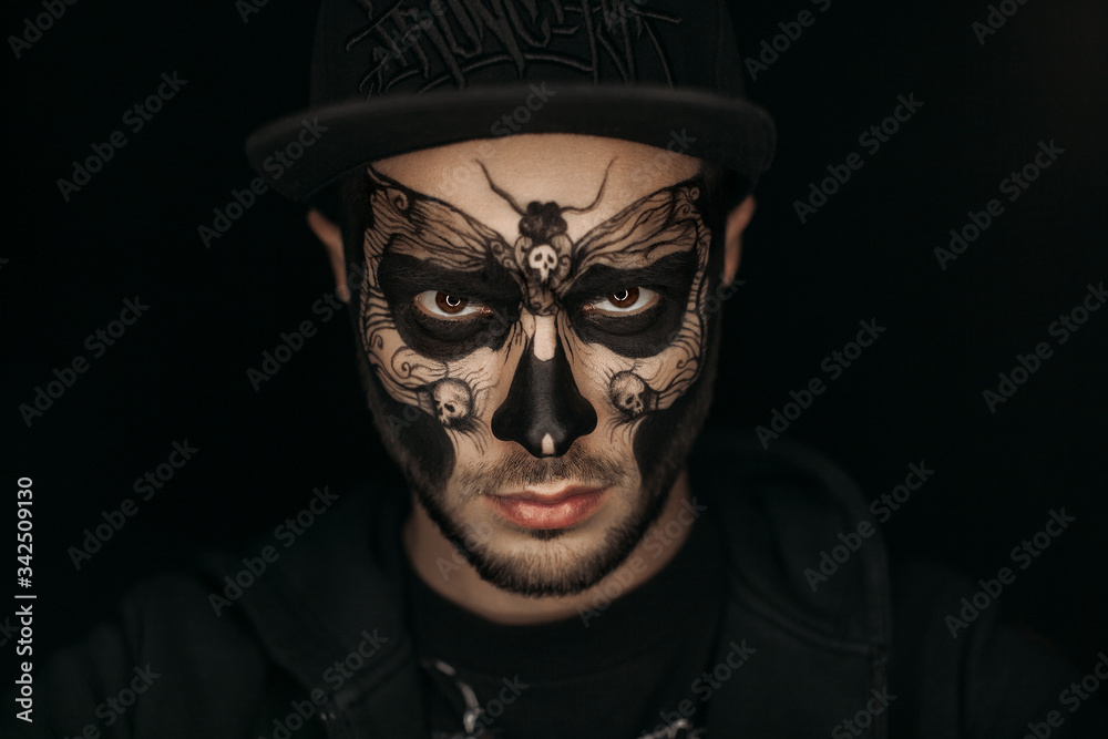 A guy with a butterfly and skulls painted on his face. Body art on the face.