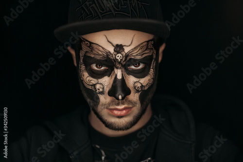 A guy with a butterfly and skulls painted on his face. Body art on the face.