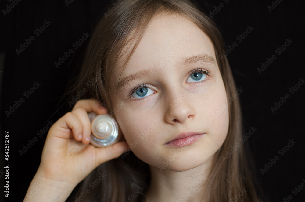 Little girl is dreaming about the summer trip to the ocean and listening to a white shell to hear a sea sound on black background in studio Little girl is dreaming about the summer trip to the ocean and listening to a white shell to hear a sea sound on black background in studio