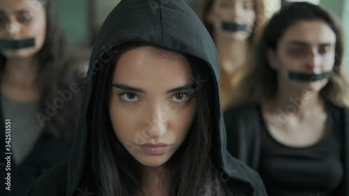 Unhappy girl in a black hood ,Sadly and depressively looking at the camera.They stand behind her,A young woman with bruises and blood on her face,