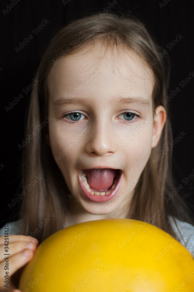Crazy funny little girl is trying to bite a big yellow pomelo in studio on black background, healthy eating concept