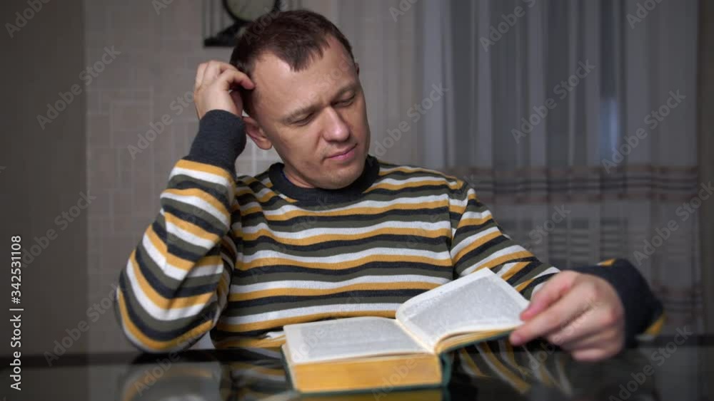 Tired young man studying the text in a book while sitting at the table in the evening Tired young man studying the text in a book while sitting at the table in the evening