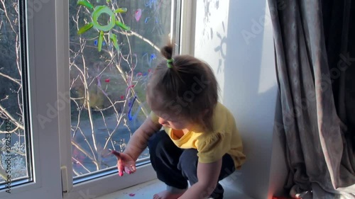 Toddler girl is painting on the window and showing her hands in the paint. Kid at home concept. Quarantine