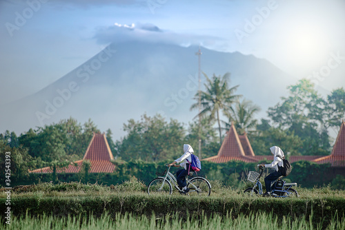 Yogyakarta Indonesia June 28, 2020 : Students on the way go to school in Sleman village, with bycyle in the morning blue sky and merapi mountain view photo