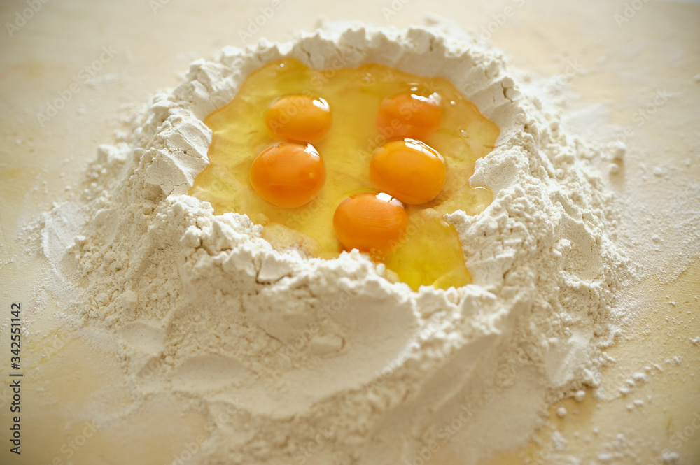 Staying at home with your family and preparing fresh home-made pasta (tagliatelle): flour on a wooden surface with a hole in the middle and eggs inside, volcano shaped. Staying at home with your family and preparing fresh home-made pasta (tagliatelle): flour on a wooden surface with a hole in the middle and eggs inside, volcano shaped.