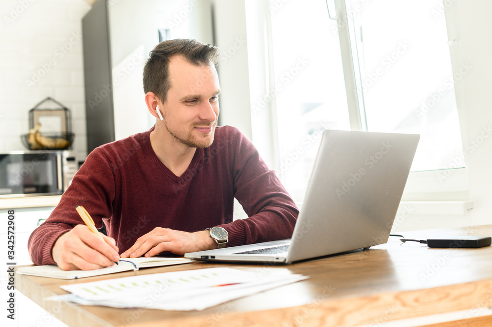 A guy uses laptop for distance study, he sits at the table at home and is writing lecture in a notebook A guy uses laptop for distance study, he sits at the table at home and is writing lecture in a notebook