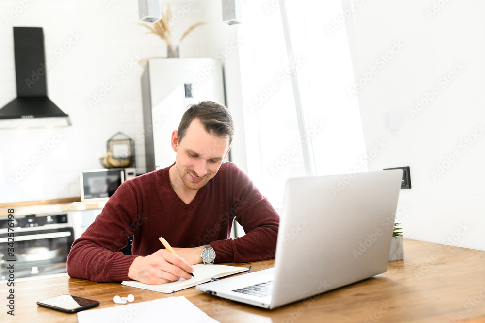 Male student writes a lecture in a notebook at home. He uses a laptop to watch a lecture. Male student writes a lecture in a notebook at home. He uses a laptop to watch a lecture.