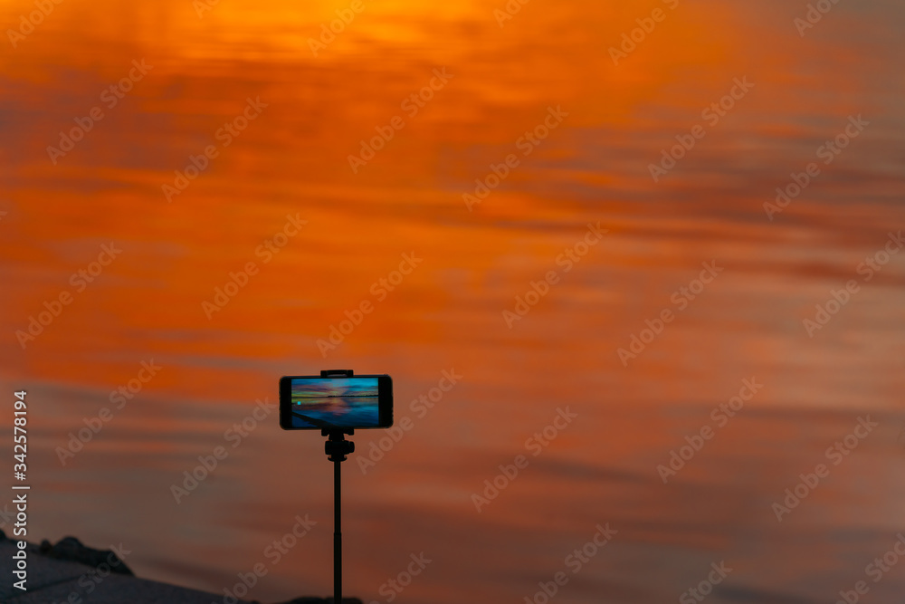 Mobile device on tripod standing by water's edge filming brilliant sunrise. Mobile device on tripod standing by water's edge filming brilliant sunrise.