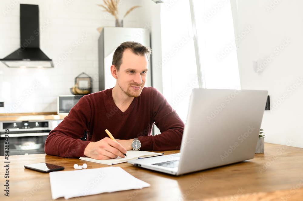 Male student writes a lecture in a notebook at home. He uses a laptop to watch a lecture. Male student writes a lecture in a notebook at home. He uses a laptop to watch a lecture.
