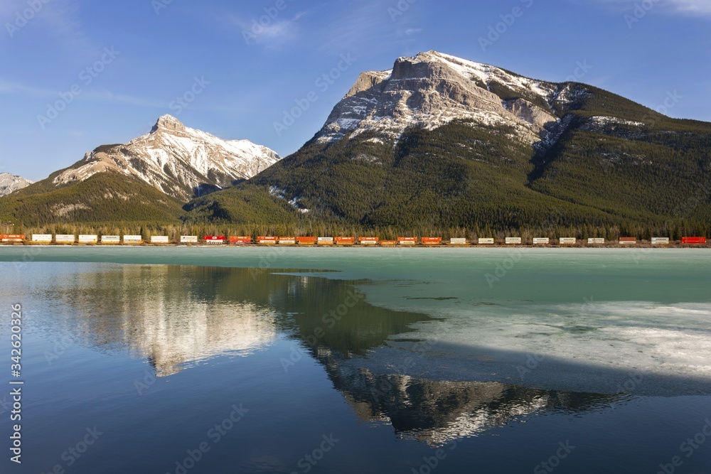 Springtime Landscape with CP Train passing through Alberta Foothills of Canadian Rockies and Snowy Mountain Peaks reflected in calm water of Gap Lake