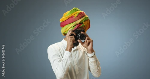 Young Hindu handsome man in traditional turban taking photos with vintage photocamera on gray wall background. Indian male making photographes with camera. photo