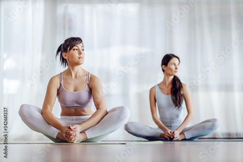 Two attractive yogi women sitting in lotus position and looking away. Yoga studio interior.